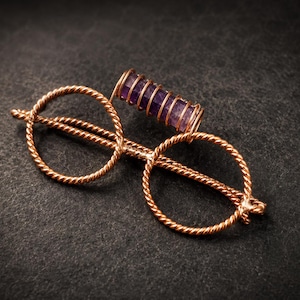 Peut inclure: Une broche en fil de cuivre en forme de lunettes rondes avec une texture métallique torsadée. Une pierre précieuse cylindrique violette est enveloppée dans un fil de cuivre fin et placée horizontalement sur le pont supérieur de la monture.