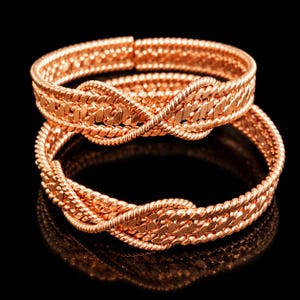Peut inclure: Deux bracelets en métal doré rose avec un motif tressé et entrelacé. Chaque bracelet est composé de plusieurs brins de fil torsadé qui se croisent pour former un motif continu et texturé avec une finition métallique polie et brillante.