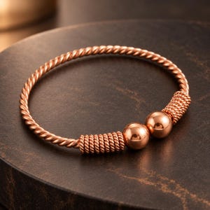 Puede incluir: Un brazalete de tono oro rosa con una banda trenzada estilo cuerda. El diseño abierto termina en dos cuentas esféricas lisas y pulidas, cada una flanqueada por una sección decorativa de alambre enrollado a lo largo de la banda metálica.