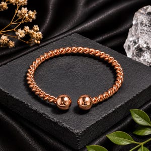 Op de afbeelding: Een roségoudkleurige manchetarmband met een gedraaide, touwachtige metalen band die eindigt in twee gladde, gepolijste bolvormige uiteinden. De armband heeft een open ontwerp voor aanpassing en een reflecterende metalen afwerking.