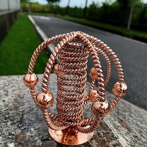 Puede incluir: Una escultura de metal color cobre que presenta una bobina vertical central rodeada por tres anillos entrelazados hechos de alambre trenzado. Cada anillo está adornado con pequeñas cuentas esféricas lisas a intervalos regulares, montadas sobre una base circular.