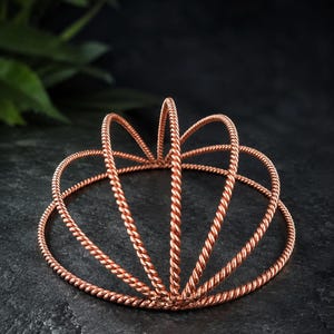 Puede incluir: Una corona o tocado de metal color cobre construido con múltiples bucles de alambre trenzado. La estructura presenta una base circular con varios segmentos de alambre arqueados que se elevan para formar un diseño simétrico y abierto.