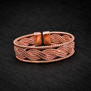 May include: A copper-colored cuff bracelet featuring a woven, braided wire design. The bracelet is constructed from multiple strands of twisted metal wire, forming a continuous pattern with two smooth, rectangular metal end caps at the opening.