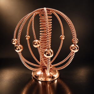 Könnte beinhalten: Eine kupferfarbene Metallskulptur mit einer zentralen vertikalen Spirale, die von mehreren konzentrischen Ringen aus gedrehtem Draht umgeben ist. Mehrere glatte, polierte kugelförmige Perlen sind an den Ringen befestigt, montiert auf einem runden Sockel.
