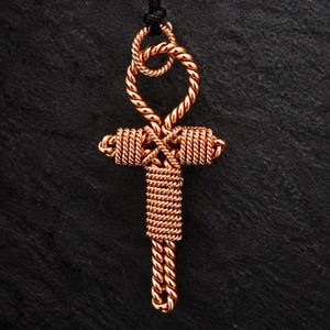 Könnte beinhalten: Ein kupferfarbener, drahtgewickelter Ankh-Anhänger mit einer gedrehten, seilartigen Textur im gesamten Design. Der Anhänger hängt an einer dünnen schwarzen Kordel und zeigt eine symmetrische Kreuzform mit einer Schlaufe oben und gewickelten Drahtdetails.