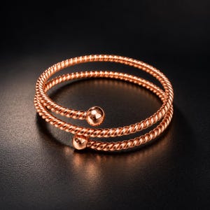 May include: A rose gold-toned bypass bracelet featuring a double-layered twisted rope design. The bracelet is constructed from two continuous strands of metallic rope, each terminating in a smooth, polished spherical bead at the open ends.