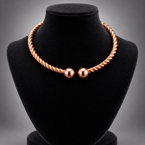May include: A copper-toned metal torque necklace featuring a twisted rope-like texture throughout the band. The necklace is an open-ended design, terminating at each end with a smooth, polished spherical ball bead of the same metallic material.