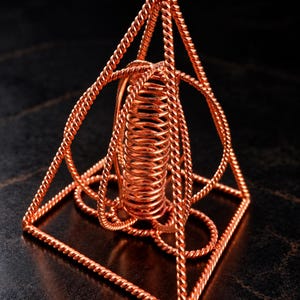 Puede incluir: Una escultura de alambre de cobre con forma de pirámide de base cuadrada. La estructura está compuesta por hebras de alambre de cobre trenzado que forman los bordes y un resorte helicoidal vertical central hecho del mismo material de cobre.