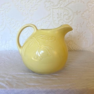May include: A pale yellow ceramic pitcher featuring a rounded body, a curved handle, and a molded floral relief design on the side. The pitcher has a wide spout and a smooth, glossy finish, showcasing a botanical motif with leaves and flower buds.