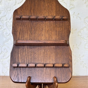 Puede incluir: Un expositor de pared de madera marrón oscuro con forma rectangular vertical y esquinas superiores redondeadas. El tablero incluye dos rieles horizontales con muescas y seis ranuras cada uno, además de un estante horizontal central sólido.