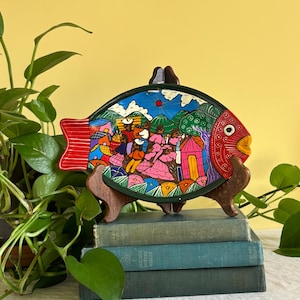 May include: A fish-shaped decorative plate featuring a hand-painted scene of people in a landscape with mountains, trees, and a red sun. The plate has a red head with a white eye, a red tail, and a dark green border, displayed on a wooden stand.