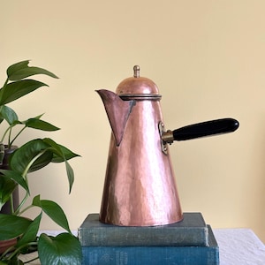 May include: A hammered copper coffee pot with a conical body, a matching lid featuring a small finial, and a black handle attached with a metal bracket. The pot has a wide base that tapers toward the top and a prominent, angular pouring spout.