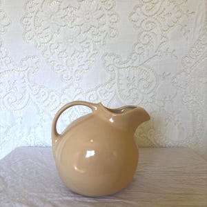 May include: A spherical ceramic pitcher with a smooth, glossy light beige finish. The vessel features a rounded body, a curved handle attached to the upper side, and a prominent pouring spout extending from the top rim of the container.