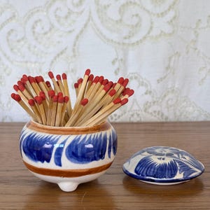 May include: A small, round, three-legged ceramic jar with a white base, blue painted floral patterns, and a brown horizontal stripe, filled with wooden matches featuring red tips. A matching ceramic lid with blue floral designs sits nearby.