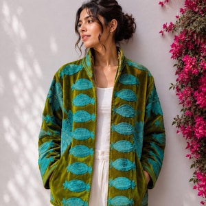 Könnte beinhalten: Eine olivgrüne Samtjacke mit einem sich wiederholenden Muster aus gestickten türkisfarbenen Fischmotiven. Die Jacke hat einen hohen Kragen, lange Ärmel und ein offenes Vorderteil, getragen über einem weißen Tanktop und einer weißen Hose.