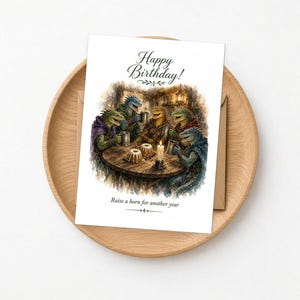 Op de afbeelding: Een rechthoekige wenskaart met een aquarelillustratie van vier hagedisachtige wezens die rond een ronde houten tafel zitten met mokken en kleine taartjes. De tekst luidt Happy Birthday! en Raise a horn for another year.