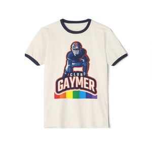 Könnte beinhalten: Ein cremefarbenes Ringer-T-Shirt mit dunkelblauem Besatz an Kragen und Ärmeln. Auf der Brust befindet sich eine Grafik eines Footballspielers in geduckter Haltung über dem Text CLUB GAYMER in Fettdruck, platziert auf einem Regenbogenstreifen.
