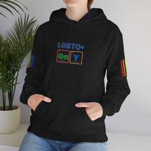 Puede incluir: Una sudadera con capucha negra que presenta un gráfico frontal con el texto LGBTQ+ y la palabra GAY estilizada como elementos de la tabla periódica Ga y Y. Las mangas tienen rayas verticales de arcoíris en los brazos exteriores.