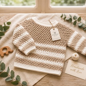 May include: A long-sleeved crochet sweater featuring horizontal stripes in beige and white. The sweater has a textured stitch pattern, a ribbed neckline, and a tag attached with string that reads Woolly Wonders Handmade. A card reads Handmade with Love.