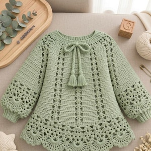 Peut inclure: Un pull à manches longues en crochet vert sauge présentant un motif ajouré complexe, une encolure ronde avec un lien décoratif se terminant par deux pompons et un ourlet festonné. Les manches présentent un large motif en crochet.
