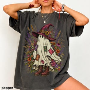 May include: A charcoal grey t-shirt featuring a graphic of a ghost wearing a purple witch hat adorned with sunflowers and wheat. The ghost is depicted with colourful patchwork squares on its body and is wearing brown lace-up boots, surrounded by wildflowers.