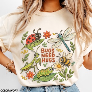 May include: An ivory t-shirt featuring a colourful graphic print of a ladybird, dragonfly, praying mantis, bee, and caterpillar surrounded by flowers and leaves. The text BUGS NEED HUGS is printed in the centre in a bold, orange-red font.