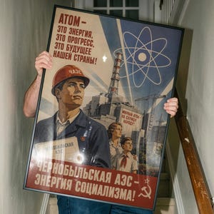 Può includere: Un poster incorniciato con un'illustrazione in stile sovietico che mostra un lavoratore con un casco rosso, una donna e un bambino davanti a una centrale nucleare. Il testo recita: ATOM - ETO ENERGIYA, ETO PROGRESS, ETO BUDUSHCHEYE NASHEY STRANY! CHERNOBYLSKAYA AES.