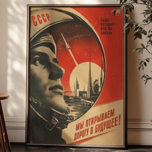 Peut inclure: Une affiche encadrée présentant une illustration de style soviétique avec un cosmonaute en casque, une fusée au décollage et une silhouette urbaine sur fond rouge. Le texte inclut CCCP, NAWE SEGODNYA - ETO IH ZAVTRA et MY OTKRYVAEM DOROGU V BUDUSCHEE.