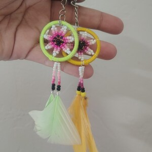May include: Two circular keychains featuring wrapped hoops in neon green and yellow. Each hoop contains a beaded star pattern in shades of pink, white, and black. Hanging from the bottom are beaded strands ending in light green and yellow feathers.