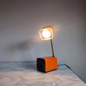 Może przedstawiać: Lampa biurkowa z jasnopomarańczową, prostokątną podstawą i pasującą kwadratową głowicą. Lampa posiada cienką, srebrną metalową szyjkę łączącą podstawę ze źródłem światła, które emituje ciepłe, jasne białe światło.