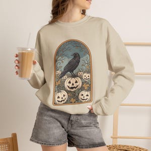 May include: A beige long-sleeve sweatshirt featuring a central graphic print of a black raven perched atop a carved jack-o'-lantern surrounded by smaller pumpkins, autumn leaves, and bare tree branches within an arched orange frame.