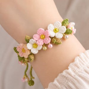 Könnte beinhalten: Ein Häkelarmband mit kleinen rosa, pfirsichfarbenen und weißen Blüten mit gelben Mitten, durchsetzt mit grünen gehäkelten Blättern und kleinen runden Perlen in Rosa- und Cremetönen, abgerundet mit herabhängenden grünen Häkelkordeln und Perlen.