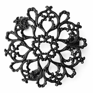 Peut inclure: Un dessous de plat circulaire en fonte noire présentant un motif ajouré complexe et symétrique composé de volutes et de formes rappelant des feuilles rayonnant depuis un centre. Le design forme une structure florale décorative.