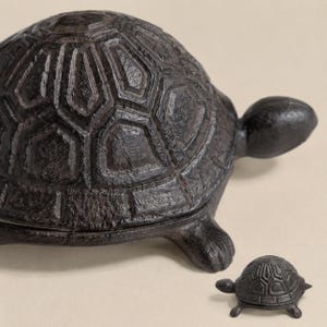 Könnte beinhalten: Eine dunkelbraune, texturierte Schildkrötenfigur aus Gusseisen mit einem detaillierten, geometrischen Panzer-Muster. Neben der größeren Schildkröte befindet sich eine deutlich kleinere, passende Schildkrötenfigur aus Gusseisen.