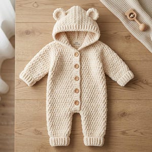 May include: A cream-colored, long-sleeved, hooded knit romper featuring a textured, chunky yarn pattern. The garment includes a hood with two small bear-like ears, five light brown buttons down the center front, and ribbed cuffs at the wrists and ankles.
