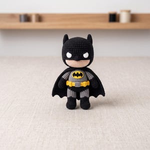 Peut inclure: Une poupée Batman au crochet avec une capuche, une cape et des bottes noires, un torse gris et une ceinture utilitaire jaune. La figurine a des yeux blancs, un visage beige et un emblème de chauve-souris noir et jaune sur la poitrine.