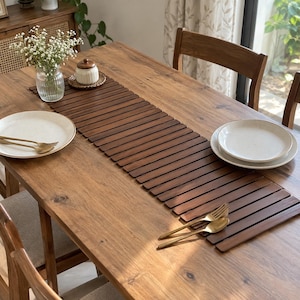 Puede incluir: Un camino de mesa rectangular de madera compuesto por listones finos de color marrón oscuro dispuestos horizontalmente. El camino está centrado en una mesa de comedor de madera, acompañado de dos platos de cerámica blanca, cubiertos dorados y un florero de cristal.