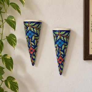 May include: Two cone-shaped wall planters featuring a vibrant blue background with hand-painted floral patterns in red, light blue, and green. Each planter has a wide, flat top rim and tapers to a point at the bottom, designed for wall mounting.