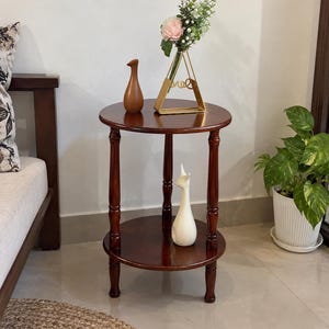 May include: A round, two-tiered wooden side table with a dark brown finish and turned legs. The top surface holds a brown ceramic vase and a gold-toned triangular frame vase with flowers. A white ceramic animal figurine sits on the lower shelf.