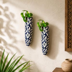 Puede incluir: Dos maceteros de pared cilíndricos con un esmalte azul profundo y un patrón floral y de enredaderas pintado a mano en blanco. Cada recipiente de cerámica contiene una pequeña planta verde, creando una exhibición vertical en la pared.