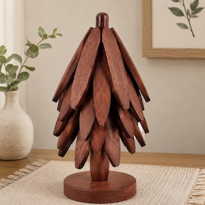 Puede incluir: Una escultura decorativa de árbol para mesa hecha de madera de color marrón oscuro. La pieza presenta un poste central vertical con múltiples listones de madera superpuestos en forma de pétalo, dispuestos para imitar un pino, sobre una base circular.