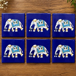 May include: A set of six square coasters with a deep blue background. Each coaster features a white elephant illustration adorned with a blue and gold decorative blanket, gold anklets, and a small floral detail on the back, all rendered in a flat style.