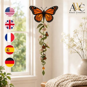 Puede incluir: Un móvil colgante que presenta una gran mariposa monarca naranja y negra en la parte superior, conectada a una enredadera verde en espiral adornada con pequeños racimos de flores rosas, tres mariposas monarca más pequeñas y una cuenta ámbar.