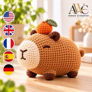 May include: A brown crochet capybara plush toy with a cream-colored snout, dark brown ears and feet, and a small orange fruit with a green leaf resting on its head. The toy features embroidered eyes and a nose, with a textured yarn construction.
