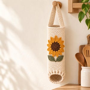 May include: A cylindrical, cream-colored crochet bag designed for hanging storage. The front features a prominent, textured sunflower appliqué with a dark brown center, bright yellow petals, and two green leaves attached near the base.