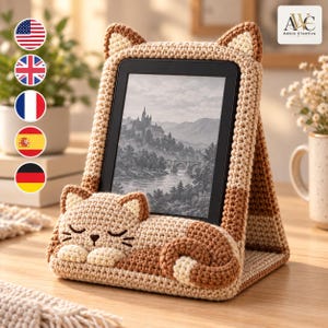 May include: A crochet tablet or e-reader stand featuring a tan and brown cat design. The stand has a sleeping cat figure at the base with ears at the top corners. The screen displays a black and white landscape image of a castle and a bridge.