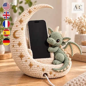 Puede incluir: Un soporte para teléfono de ganchillo color crema con forma de media luna, decorado con estrellas doradas bordadas. Un pequeño dragón de ganchillo verde salvia, con alas y cuernos color crema, está sentado en la base sosteniendo un smartphone negro.