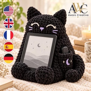 May include: A black crochet cat tablet stand featuring a smiling face with lavender embroidered whiskers and nose. The cat holds a black e-reader displaying a moon and stars graphic. A small side pocket features a lavender crescent moon embroidery.