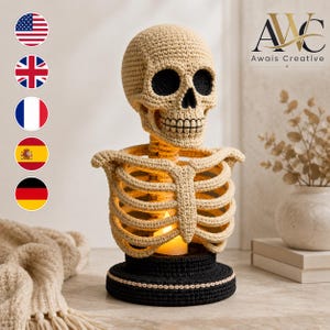 May include: A crocheted beige skull and rib cage sculpture mounted on a circular black base. The hollow chest cavity contains a glowing yellow LED candle. The piece features detailed stitching throughout the skull and rib structure.