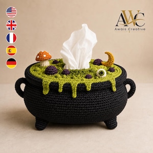 May include: A black crocheted tissue box cover shaped like a cauldron with a green textured top featuring a white tissue, a small orange mushroom, a yellow crescent moon, and a single white eyeball with a green iris and black pupil.